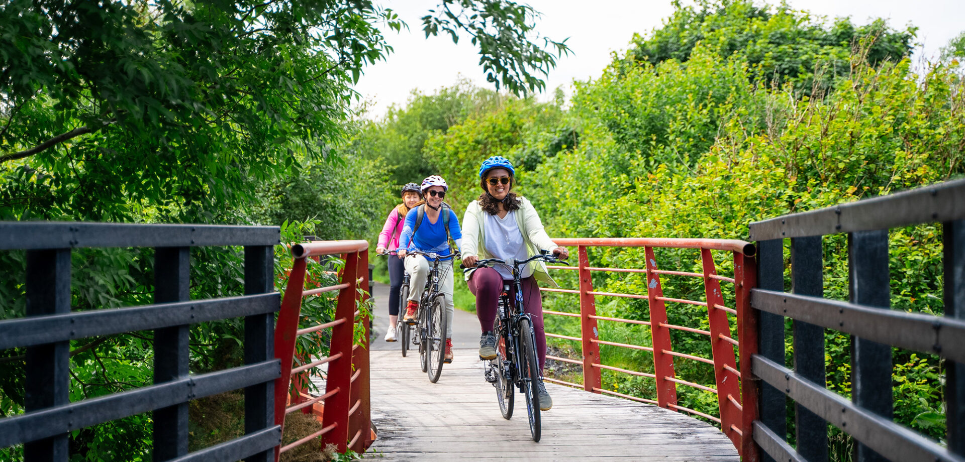 Three people wearing helmets ride bicycles across a wooden bridge surrounded by lush green trees and vegetation.