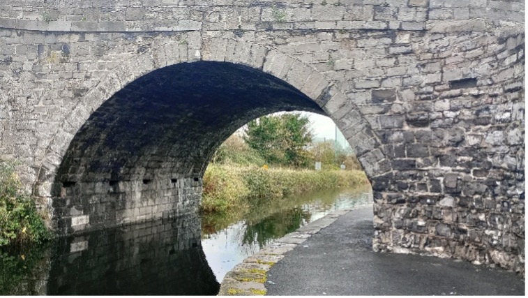 Photograph of a stone arch bridge over a narrow canal with a paved towpath running alongside water. The towpath is noticeably narrow under the bridge The scene includes reflections of the arch in the water, dense greenery, and a cloudy sky in the background.