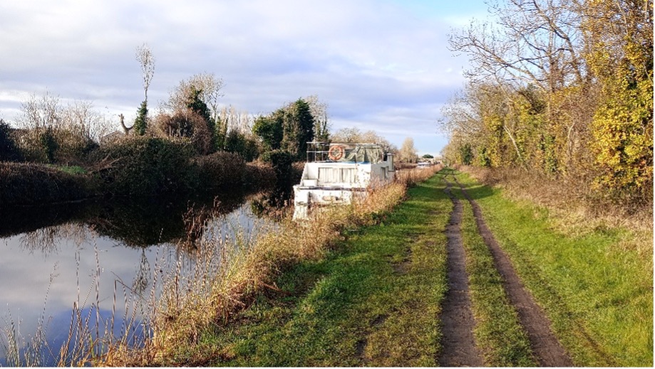 Photograph of a narrow canal with a white boat moored alongside a grassy towpath bordered by autumnal trees and bushes. The scene captures a calm, rural setting with reflections on still water and a partly cloudy sky.