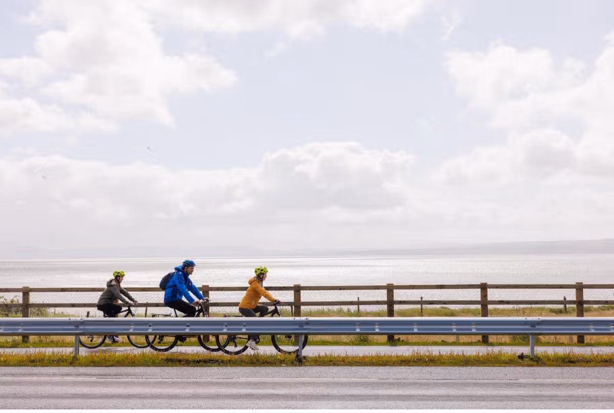 Photograph showing three cyclists riding along a coastal road with a metal guardrail separating them from a road and on the other side, a wooden fence from a body of water under a cloudy sky. Cyclists wear helmets and jackets in grey, blue, and yellow.