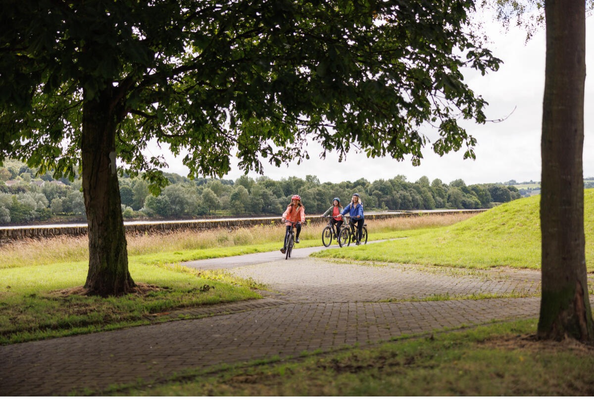 Photograph showing three people cycling on a paved path through a green park with large trees and a riverbank in the background.