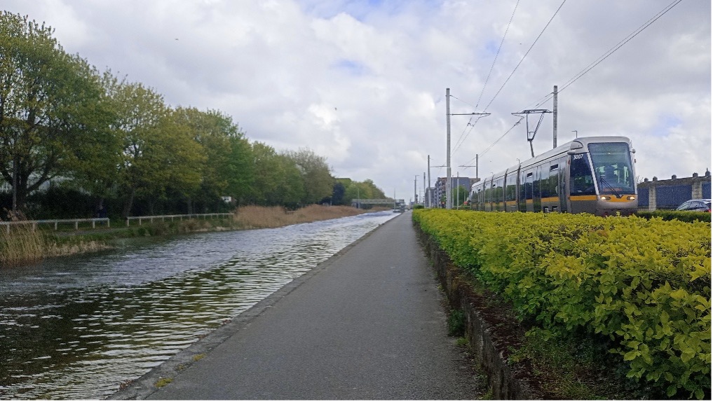 Photograph showing a tram travelling along a track beside a canal with a paved pathway in between. The scene includes green bushes on one side, trees on the other, and a cloudy sky overhead, highlighting a blend of urban transport and natural elements.