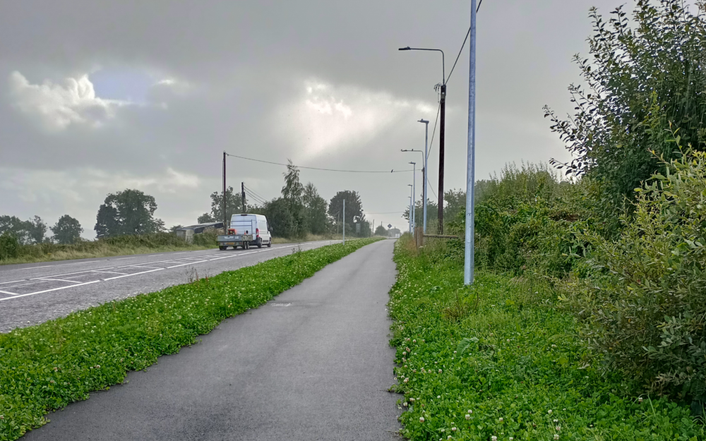 Photograph of a tarmac route running alongside a road separated by green vegetation and streetlights under a cloudy sky. The scene highlights a quiet, rural setting with a mix of natural greenery and infrastructure elements like power lines and a distant vehicle.