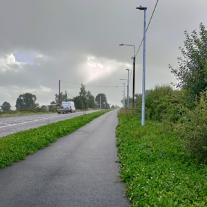 Photograph of a tarmac route running alongside a road separated by green vegetation and streetlights under a cloudy sky. The scene highlights a quiet, rural setting with a mix of natural greenery and infrastructure elements like power lines and a distant vehicle.