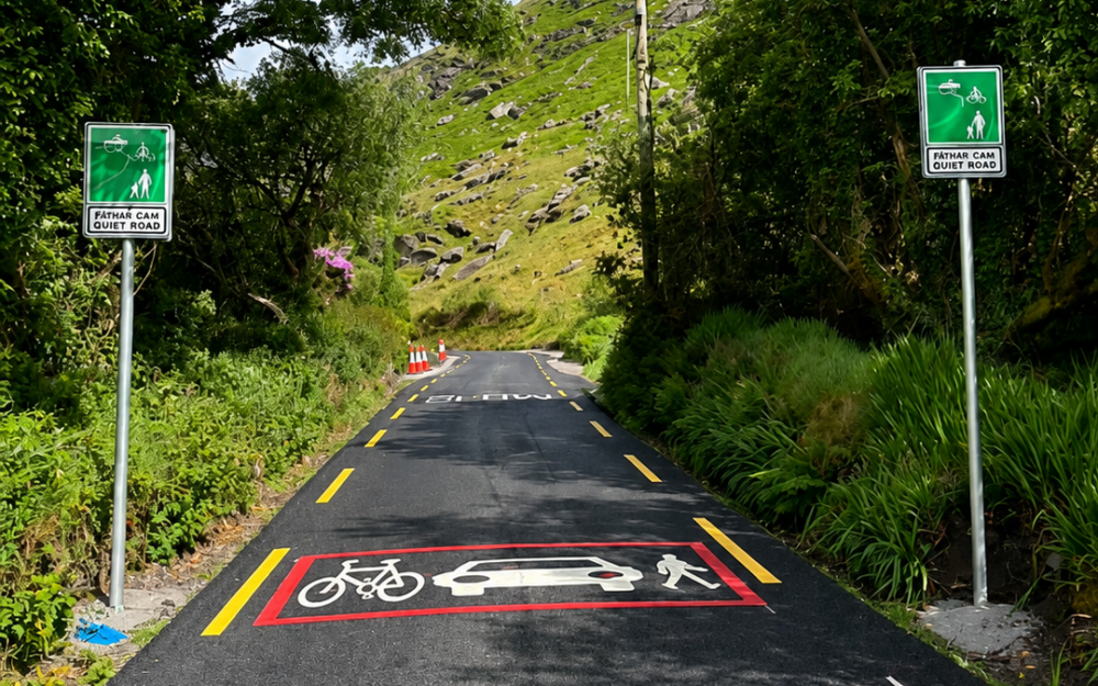 Photograph of a narrow road or path through a green, wooded area with hills in the background, featuring road markings and signs indicating a shared space for bicycles, cars, and pedestrians. Two green signs on either side of the path display a pedestrian and cyclist symbol with the text "SHARED SPACE DRIVE SLOW," while the road markings include white icons of a bicycle, car, and pedestrian within a red-bordered rectangle.