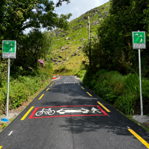 Photograph of a narrow road or path through a green, wooded area with hills in the background, featuring road markings and signs indicating a shared space for bicycles, cars, and pedestrians. Two green signs on either side of the path display a pedestrian and cyclist symbol with the text "SHARED SPACE DRIVE SLOW," while the road markings include white icons of a bicycle, car, and pedestrian within a red-bordered rectangle.